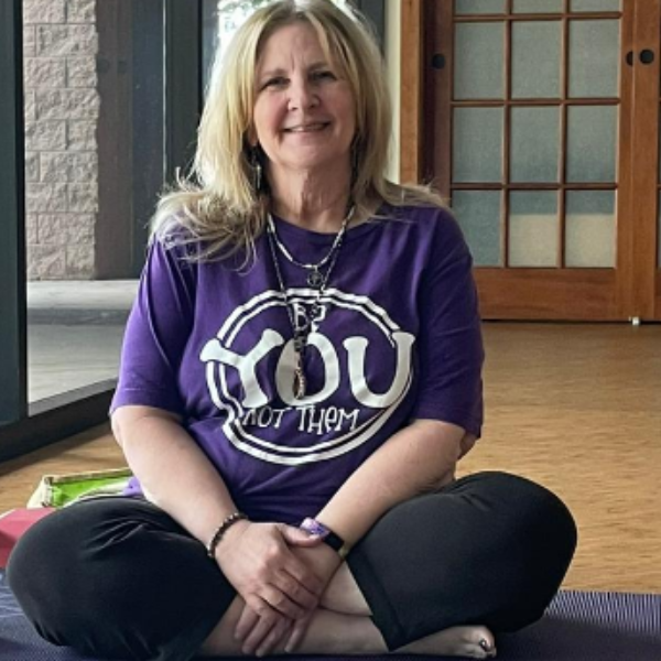 Smiling woman seated cross-legged on a yoga mat, wearing a purple ‘Be YOU not them’ shirt, radiating warmth and empowerment in a peaceful studio setting.